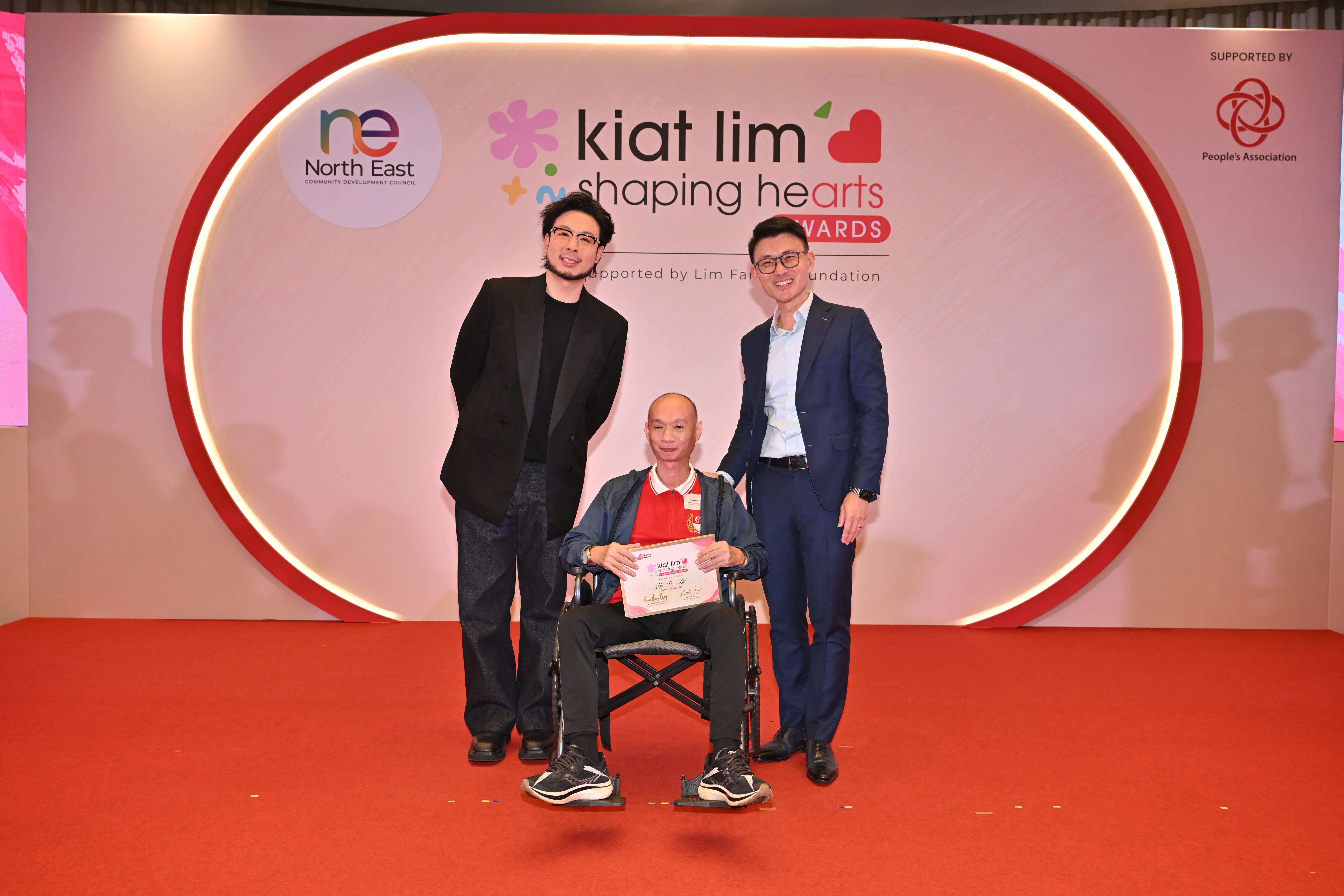 Award recipient standing on stage with the presenter, holding their certificate and posing for a group photo during the award ceremony, with a backdrop reading ‘Kiat Lim-Shaping Hearts Awards'.